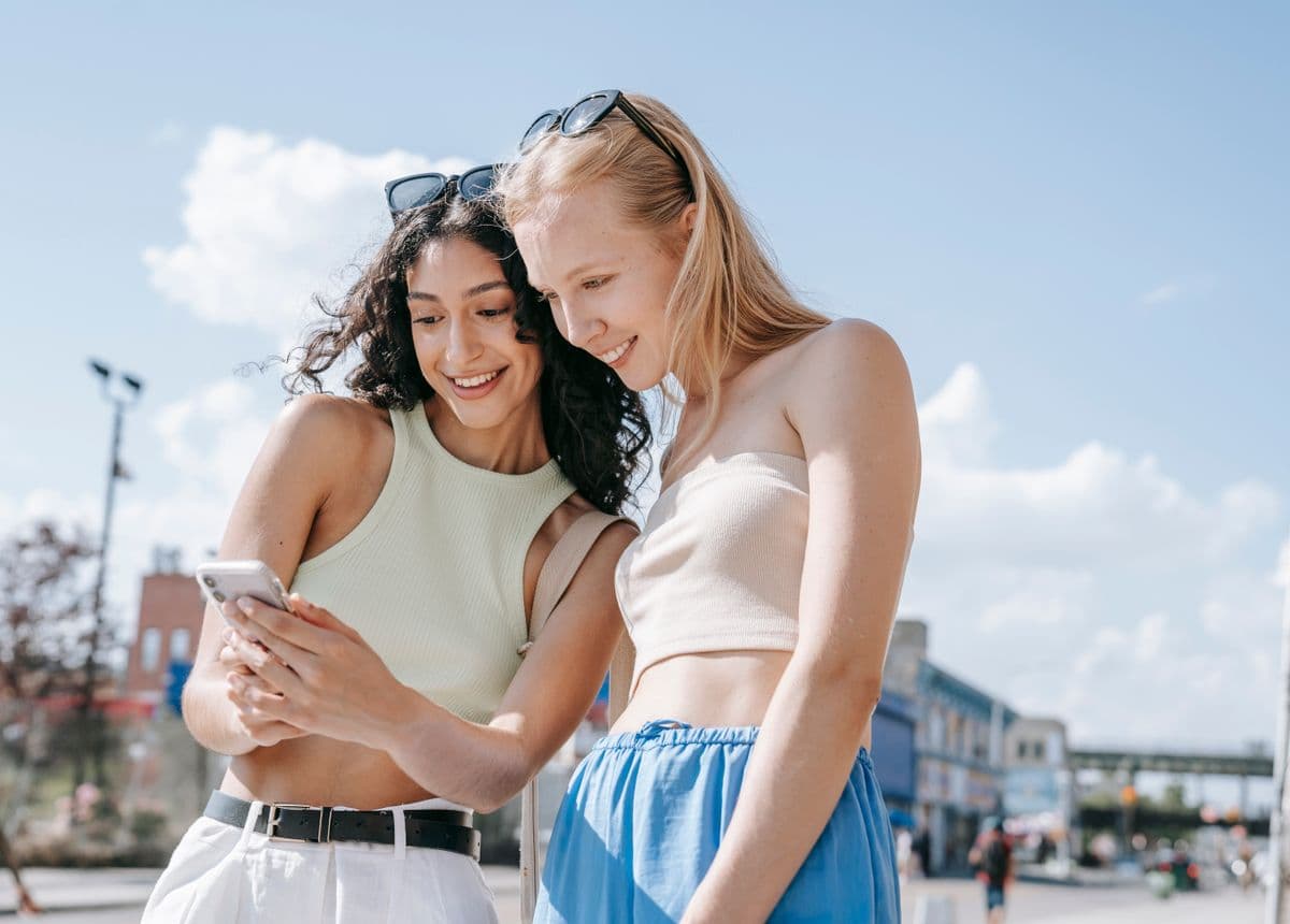 Two travellers smiling at a phone screen together under a bright blue summer sky